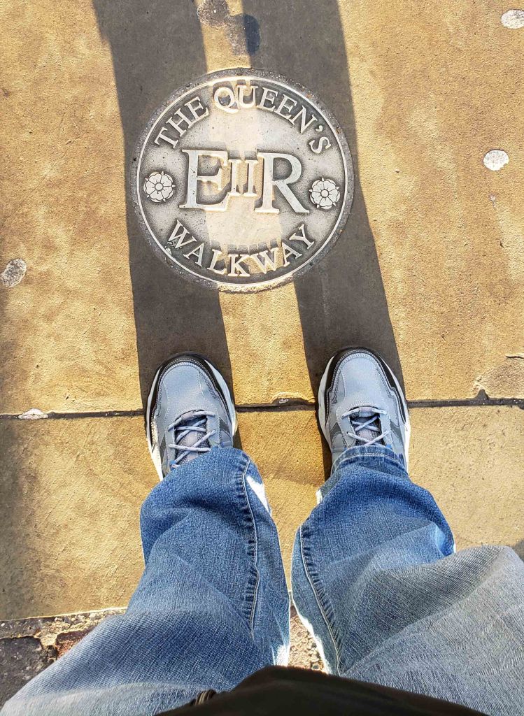 A person's feet standing on a paved walkway marked 'The Queen's Walkway' in Windsor, England.