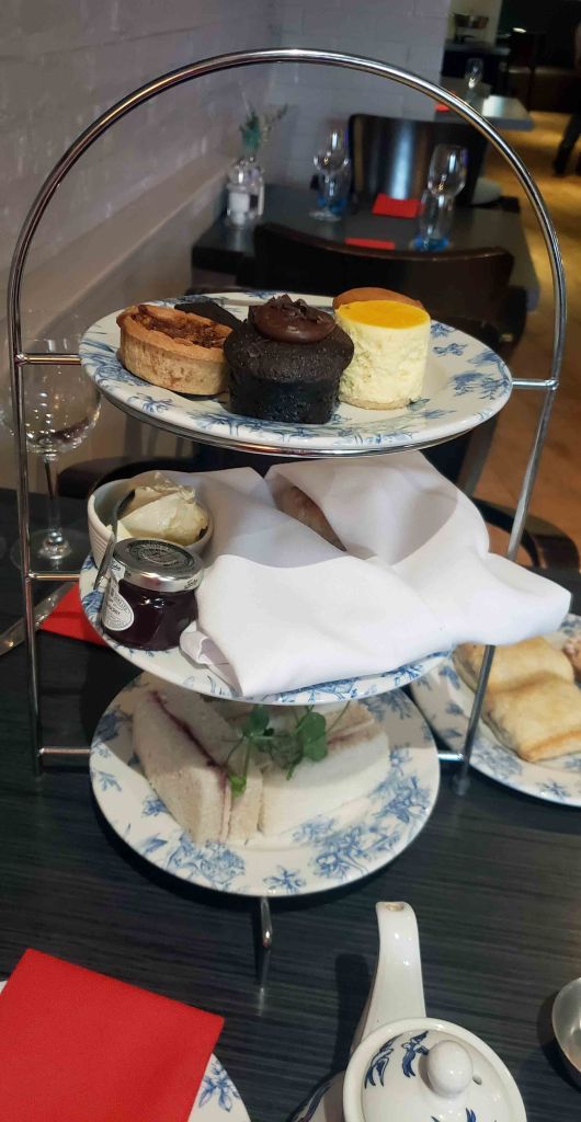 A tiered stand displaying an assortment of sweets and sandwiches for an afternoon tea setting, featuring pastries, assorted cakes, butter, and jam, alongside a teapot and red napkins on a dining table.