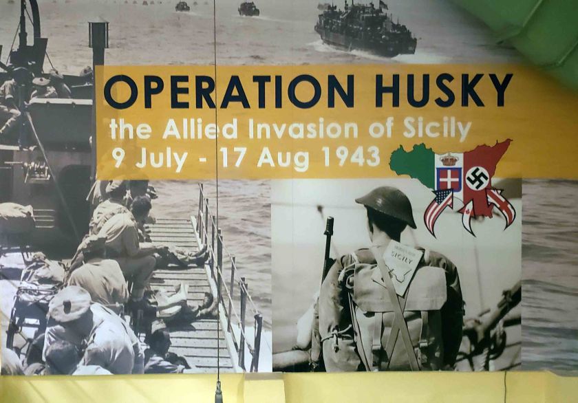 A historical display titled 'Operation Husky' highlighting the Allied invasion of Sicily from July 9 to August 17, 1943. The background features images of soldiers embarking on landing crafts and military maps with flags representing Italy and Germany.