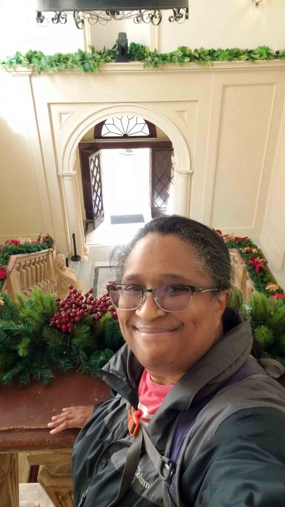 Selfie of a person with glasses smiling at the camera, standing near a staircase decorated with greenery and red berries, with an entrance door visible in the background.