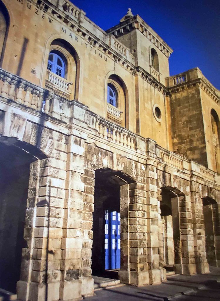 Facade of a historic stone building with arched doorways and blue doors, featuring a balcony and signage reading 'MUZEW MARITTIMU' in a clear sky.