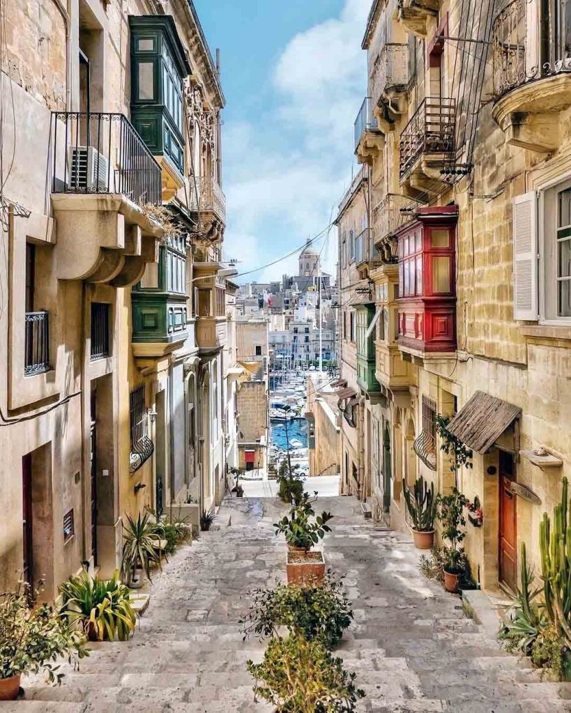 A narrow street lined with colorful balconies and potted plants, leading down towards a marina with boats in the background.